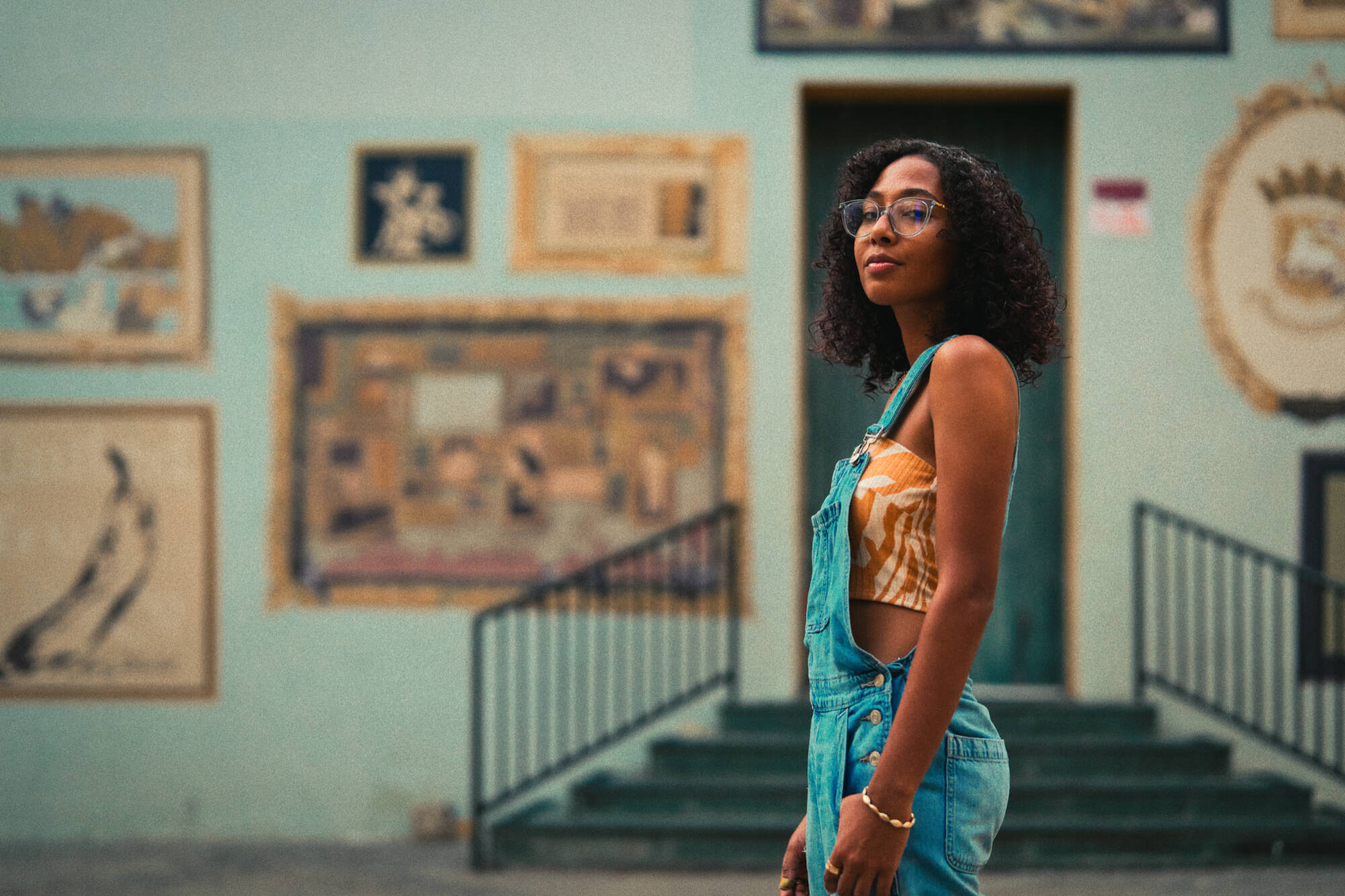 A color photograph of a person in profile from the calves-up standing in front of a building. They have medium-dark skin and curly dark hair down to their shoulders
